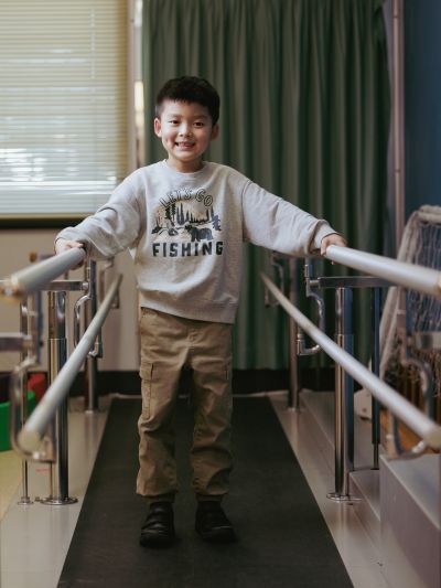 A child standing between two parallel support bars in a therapy or rehabilitation room, wearing a light grey “Let’s Go Fishing” sweatshirt and khaki pants, with curtains and a window in the background.