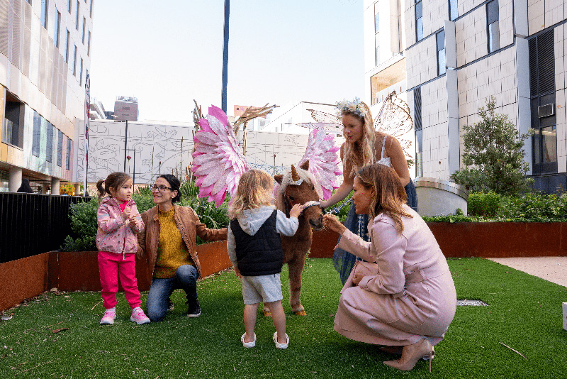 Two little kids and their mums at Sydney Sick Kids Day petting a pony wearing big pink wings
