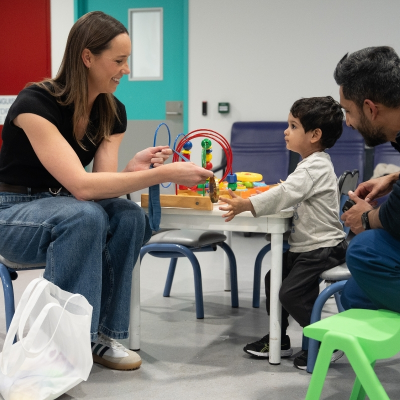 Emma Mckeon sitting down with a child in hospital, playing with toys. The boys father sits behind smiling.