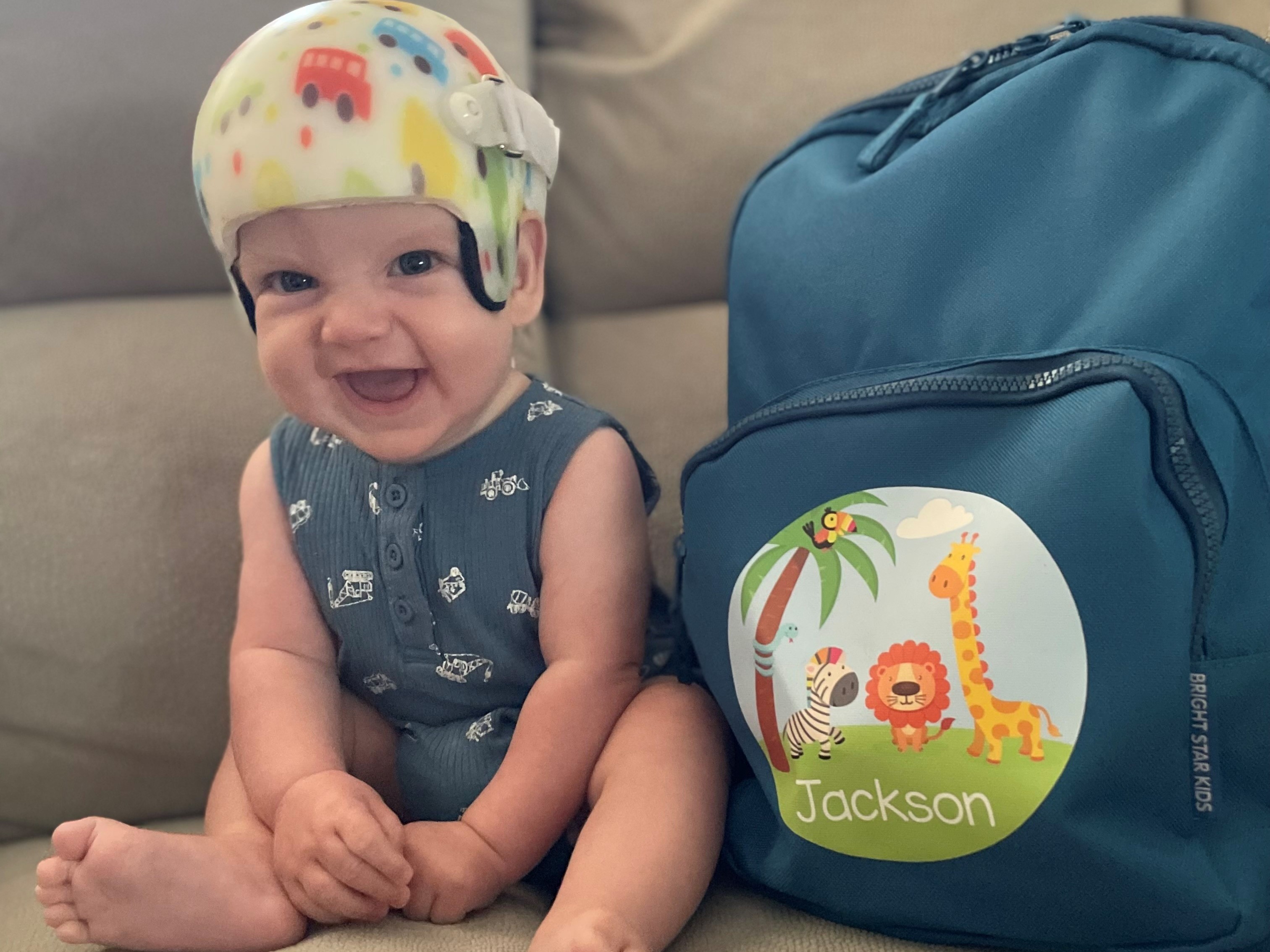 miling baby in a colorful helmet and blue romper sitting on a couch next to a blue animal-themed backpack labeled 'Jackson'. miling baby in a colorful helmet and blue romper sitting on a couch next to a blue animal-themed backpack labeled 'Jackson'.