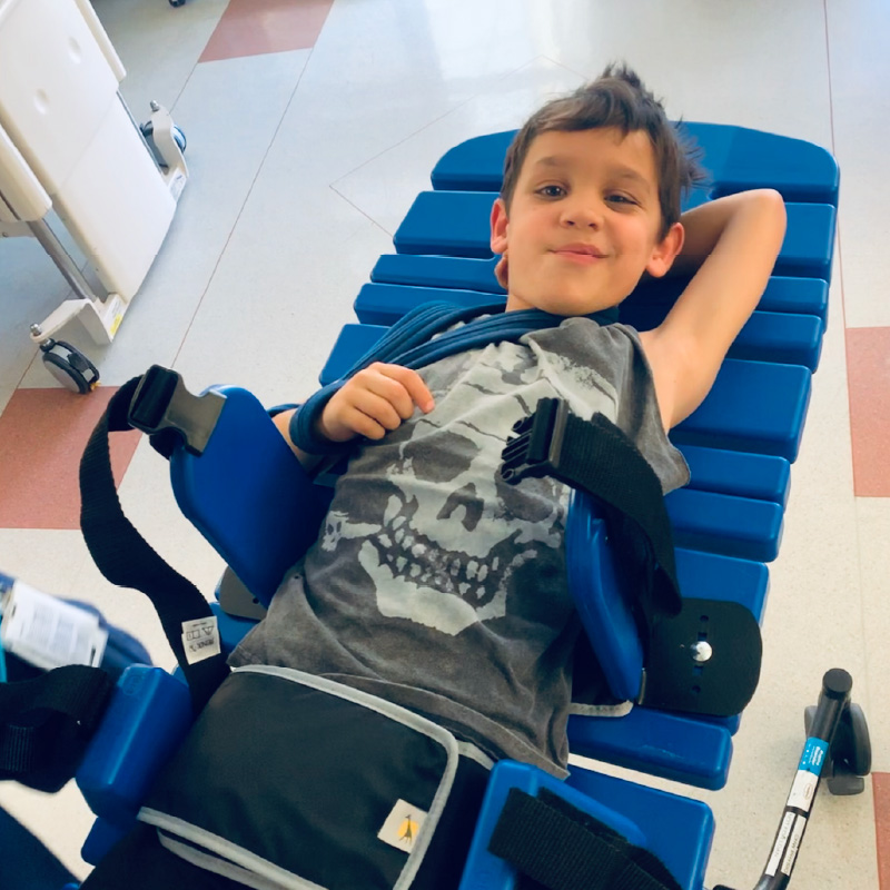 Hugo laying on a rehab bed with his right arm in a sling, his other arm resting above his head. He has a big grin on his face, enjoying himself during rehabilitation sessions.