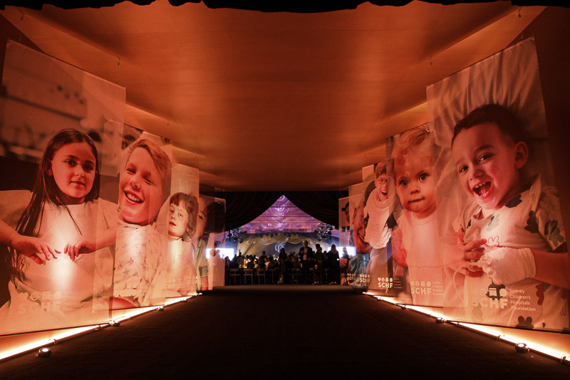 Giant posters of children in hospital line a brightly lit walkway into Gold Dinner 2025 Giant posters of children in hospital line a brightly lit walkway into Gold Dinner 2025