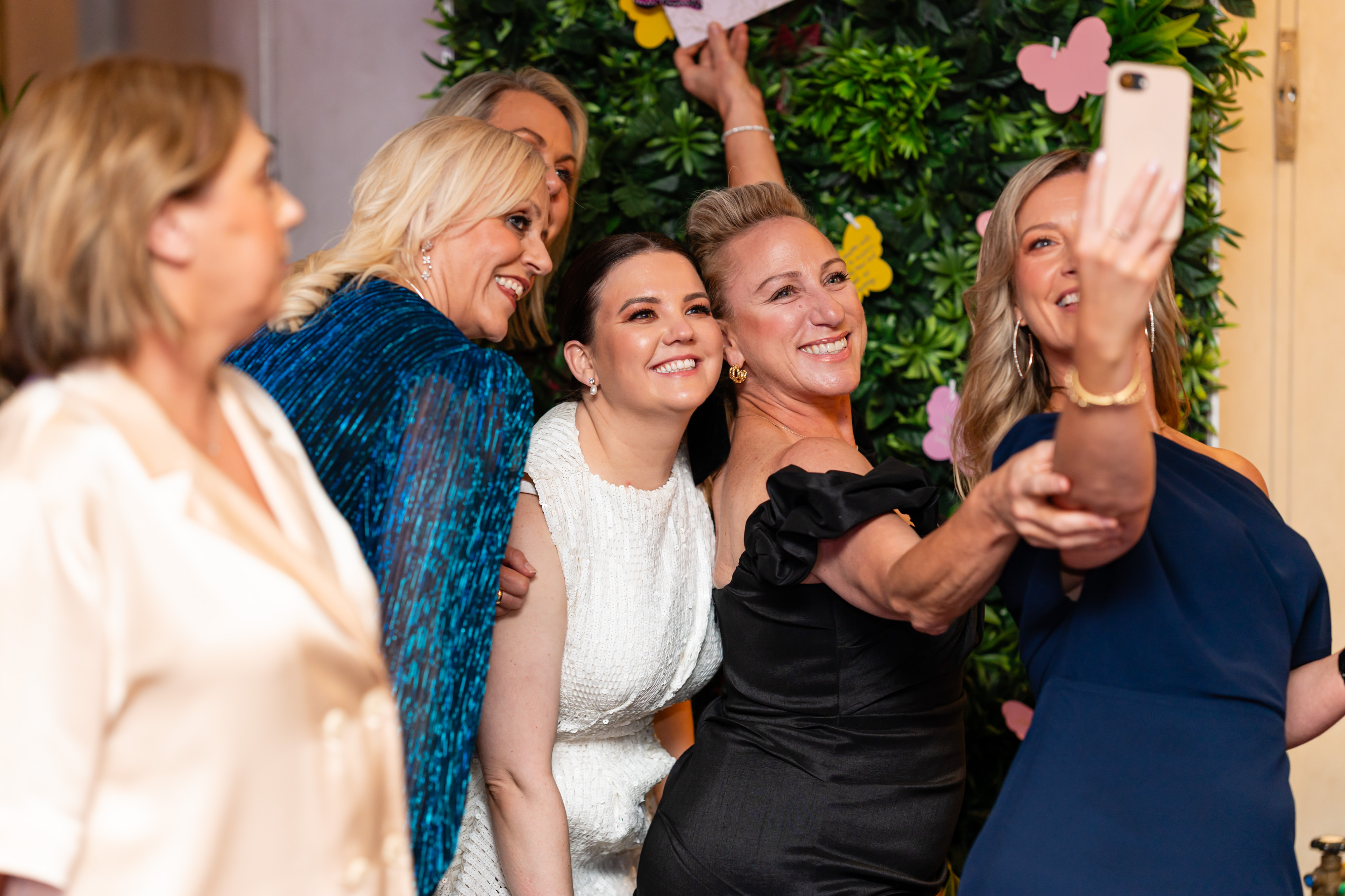 Guests in formal attire leaning together and taking a selfie in front of a green floral wall.