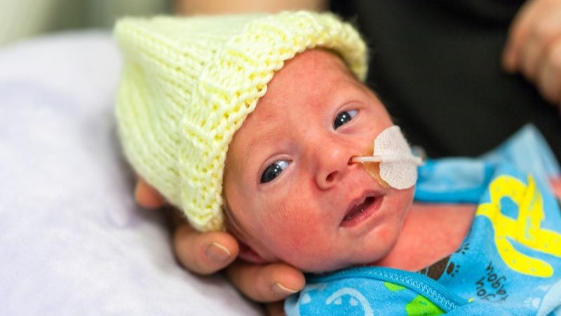 Newborn baby in hospital wearing a knitted hat, holding a caregiver’s finger