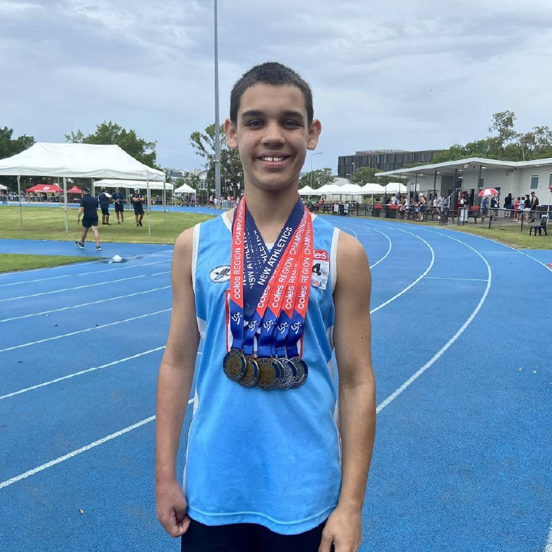 Hugo as a teen standing on an athletics track wearing a blue singlet with six medals draped around his neck.