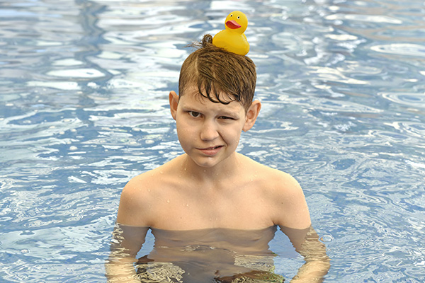 Caleb as a teenager swimming in a large pool with a yellow rubber duck resting on his head