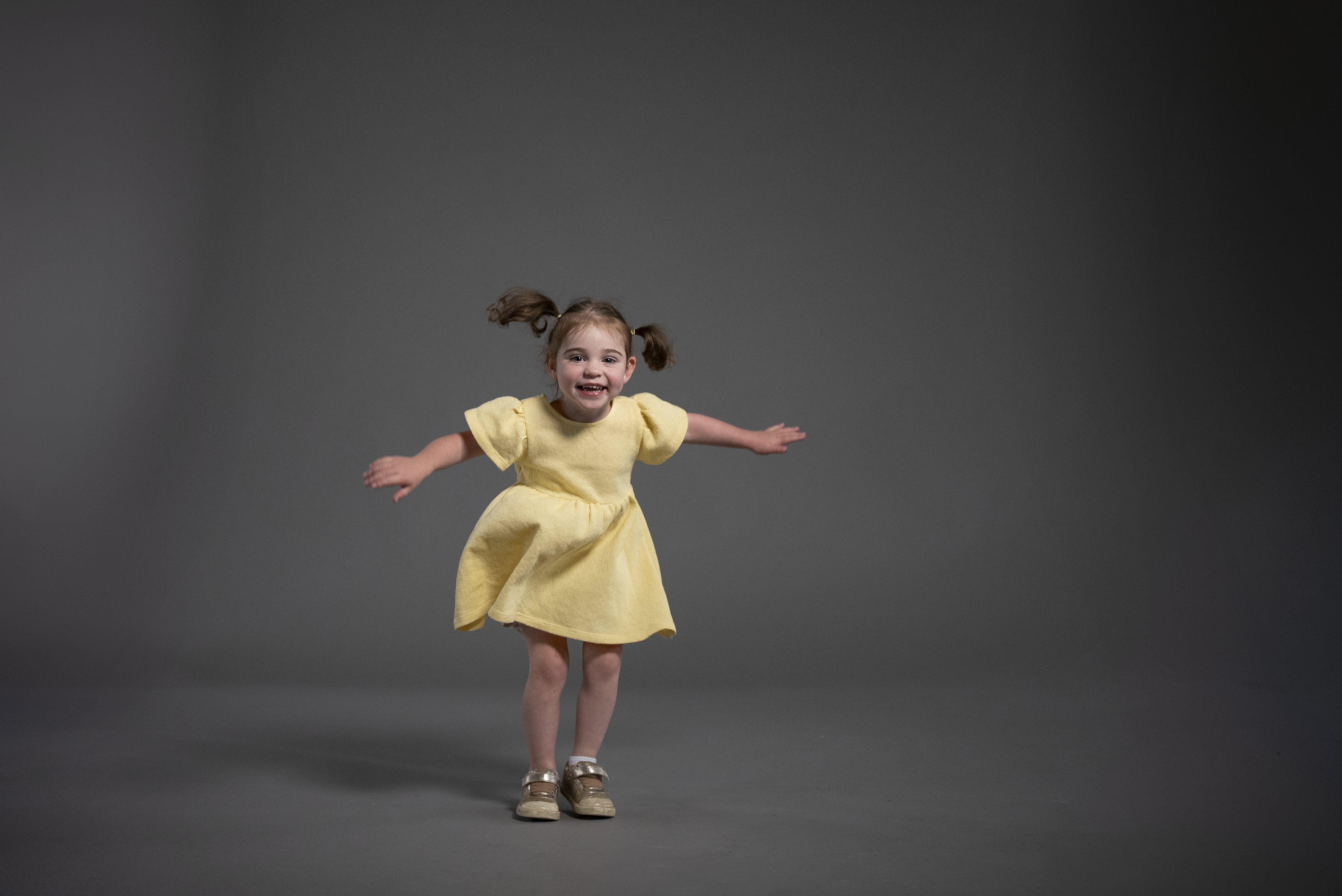Image of child wearing yellow dress and jumping with hands out with grey background.
