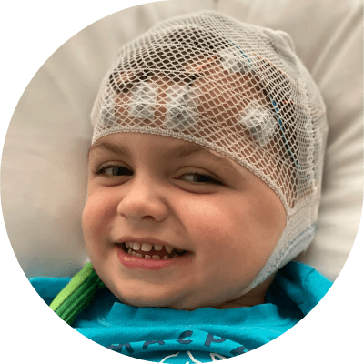 Noah lying in a hospital bed. he is wearing a cloth wrap on his head covering brain wave measuring equipment that is taped to his forehead. He's got a cheeky grin.