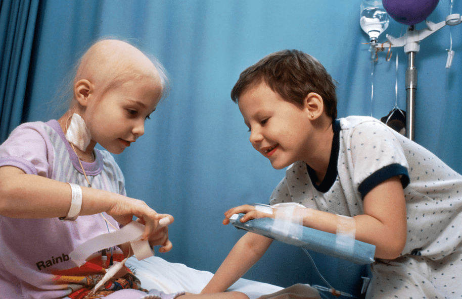 A young girl and boy in hospital gowns giggling as they play with hospital tape, helping each other wrap their arms and fingers up.