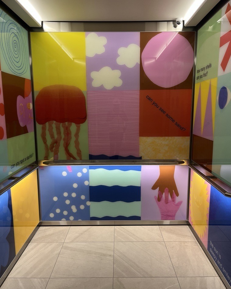Elevator interior decorated with colorful geometric panels featuring abstract shapes, clouds, jellyfish, waves, and text prompts like ‘Can you see some shapes?’ and ‘How many shells can you find?