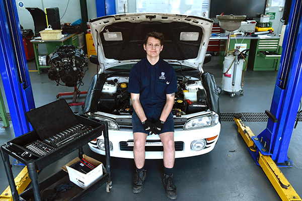 Caleb as an adult leaning on the front of a car in a mechanic workshop. He wears a mechanic outfit with a trolley full of spanners and tools next to him