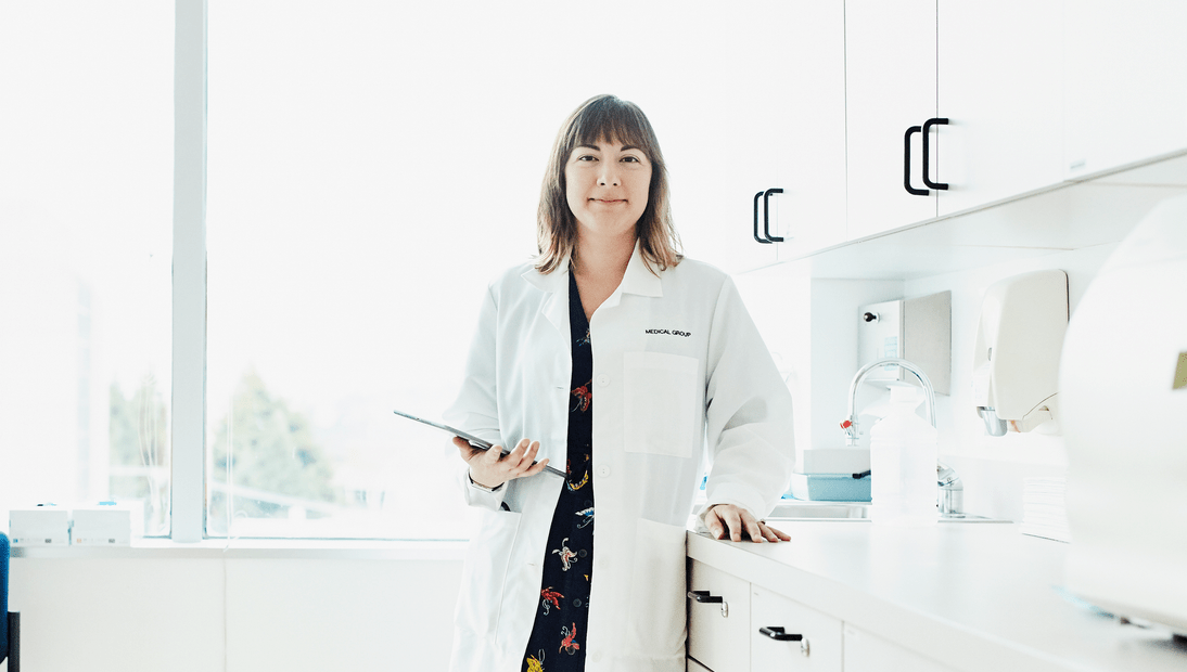 Smiling female doctor in a white coat holding a tablet, standing in a bright, modern medical clinic with natural light