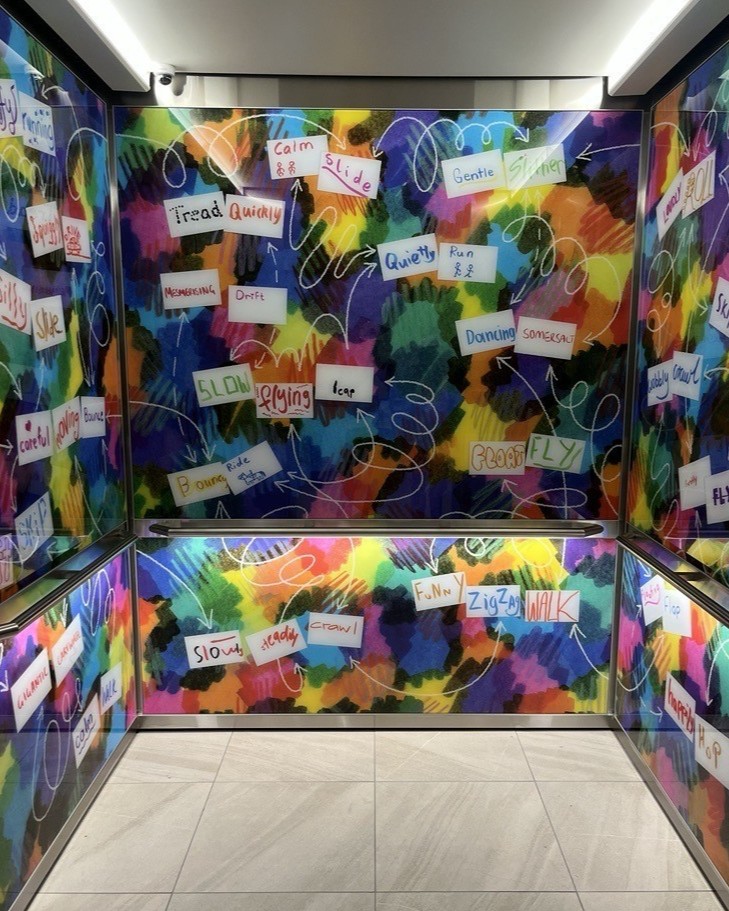 Elevator interior decorated with a vibrant, multicolored abstract background featuring handwritten words like ‘Calm,’ ‘Slide,’ ‘Slow,’ and ‘Zigzag Walk’ scattered across the walls.