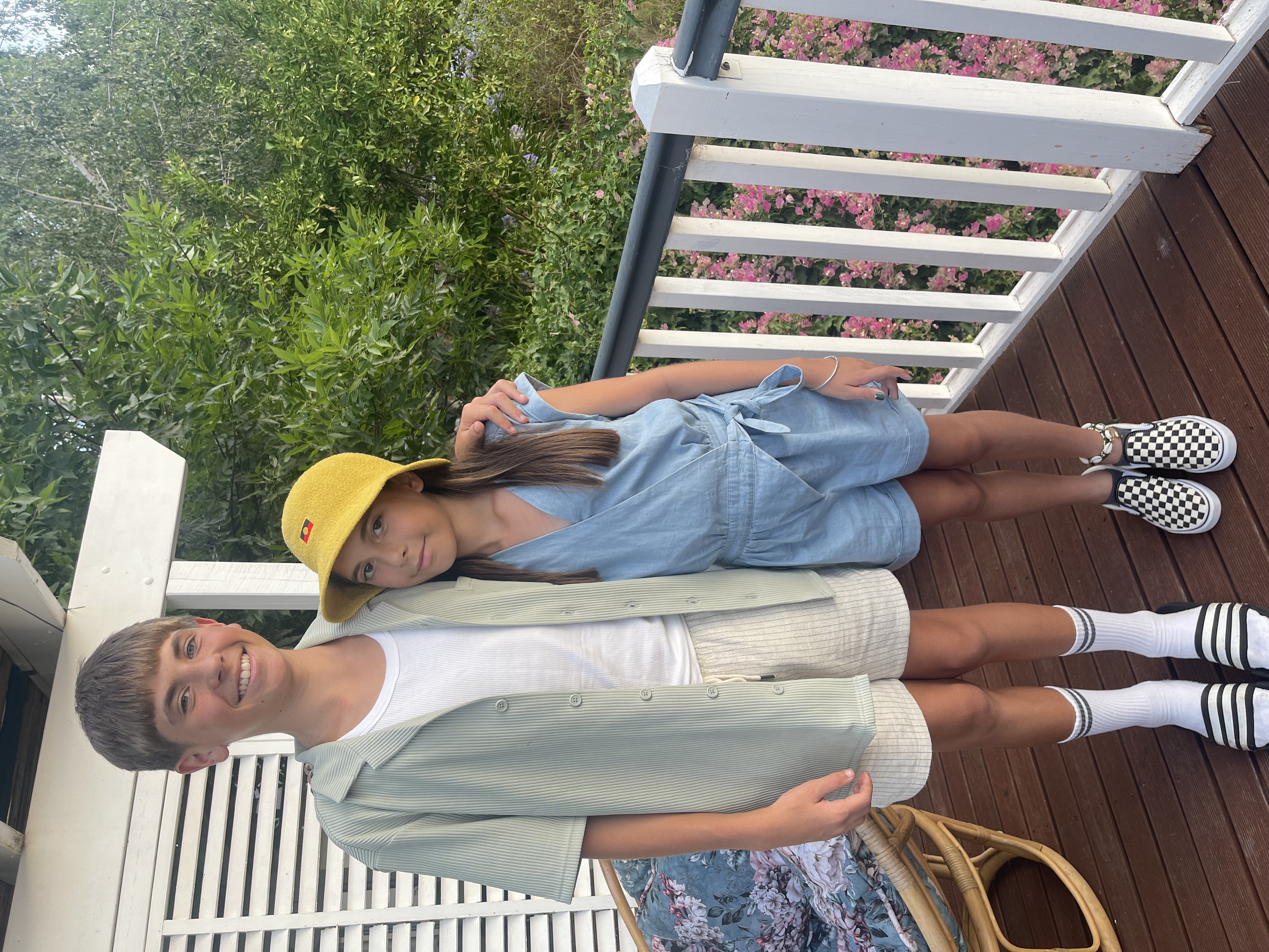 Two children standing together on a verandah outdoors, dressed in casual summer clothes.