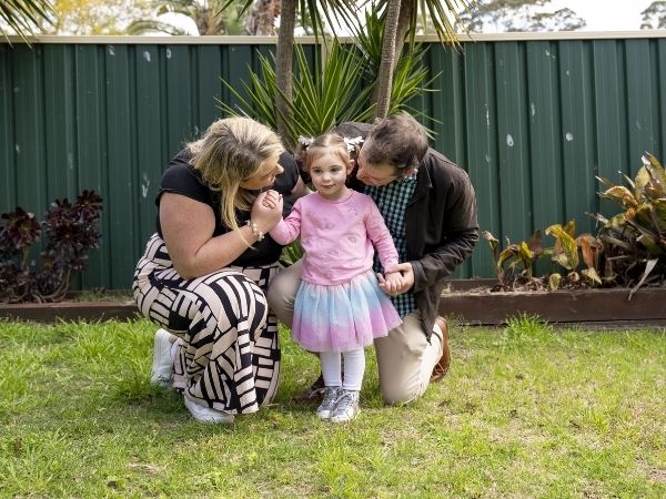A young girl and her parents holding hands in their backyard. The girl wears a bright pink jumper and a multi-coloured tutu