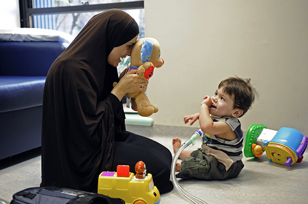 a toddler boy sitting on the floor next to his mum laughing as she holds a fluffy teddy bear in front of her face