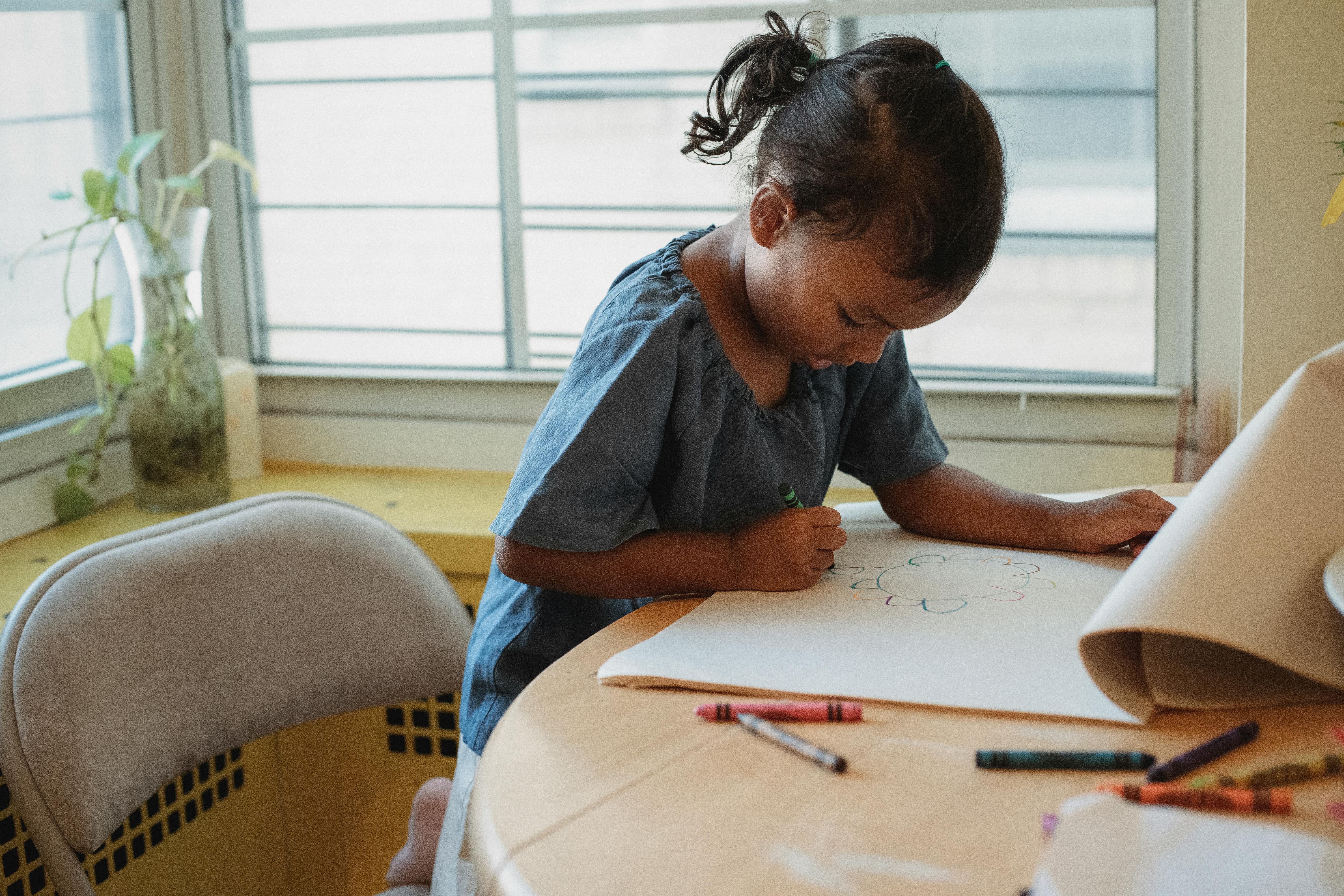Image of young girl leaning on chair with colouring pencils, doing a drawing in a sketch book.