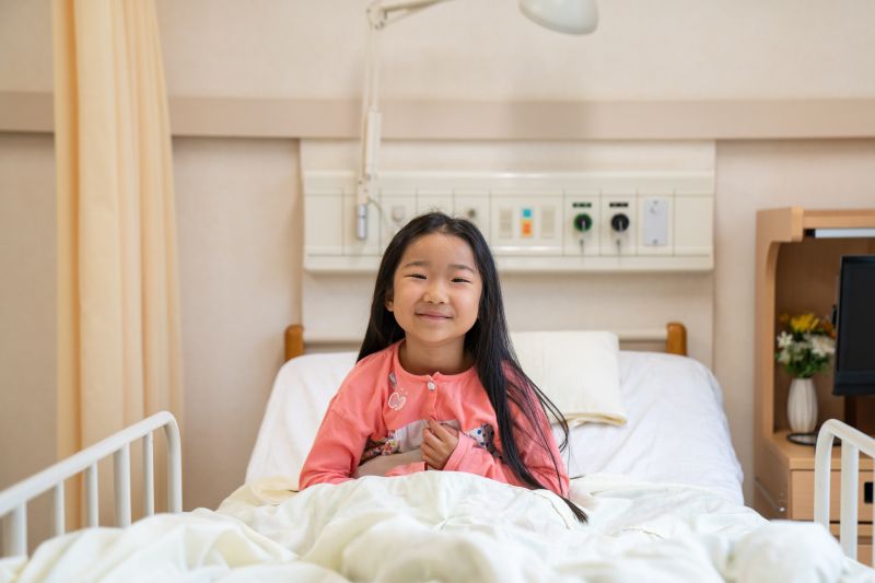 hild sitting upright in a hospital bed, surrounded by medical equipment and a bedside table with flowers.