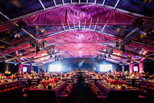 The interior of the event space at Gold Dinner 2025. A large open dining room with many long, lavishly decorated tables in SCHF purple colours. Bright purple banners above are lit by lights showing the Gold Dinner logo.