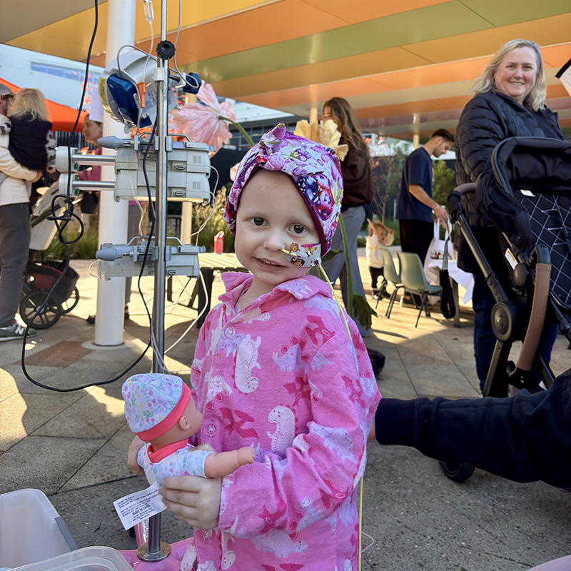 A young girl wearing bright pink pajamas and a multi-coloured beanie holding a toy baby. The girl has a breathing tube coming out of her nose, hooked up to a medical device behind her.