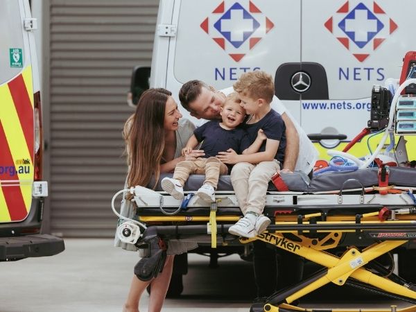 Two parents and their two young boys smiling and hugging. The two boys sit on a hospital stretcher, the older one supporting his younger brother. Behind the family two NETS ambulance vehicles can be see.