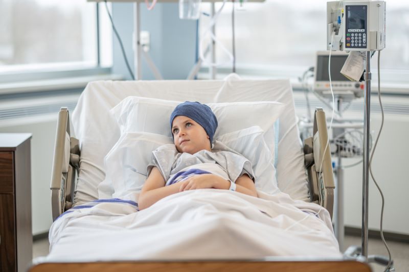A young child lying in a hospital bed, wearing a headscarf and hospital gown, looking off thoughtfully. Medical equipment and an IV pole are visible beside the bed, indicating the child is receiving treatment. A young child lying in a hospital bed, wearing a headscarf and hospital gown, looking off thoughtfully. Medical equipment and an IV pole are visible beside the bed, indicating the child is receiving treatment.