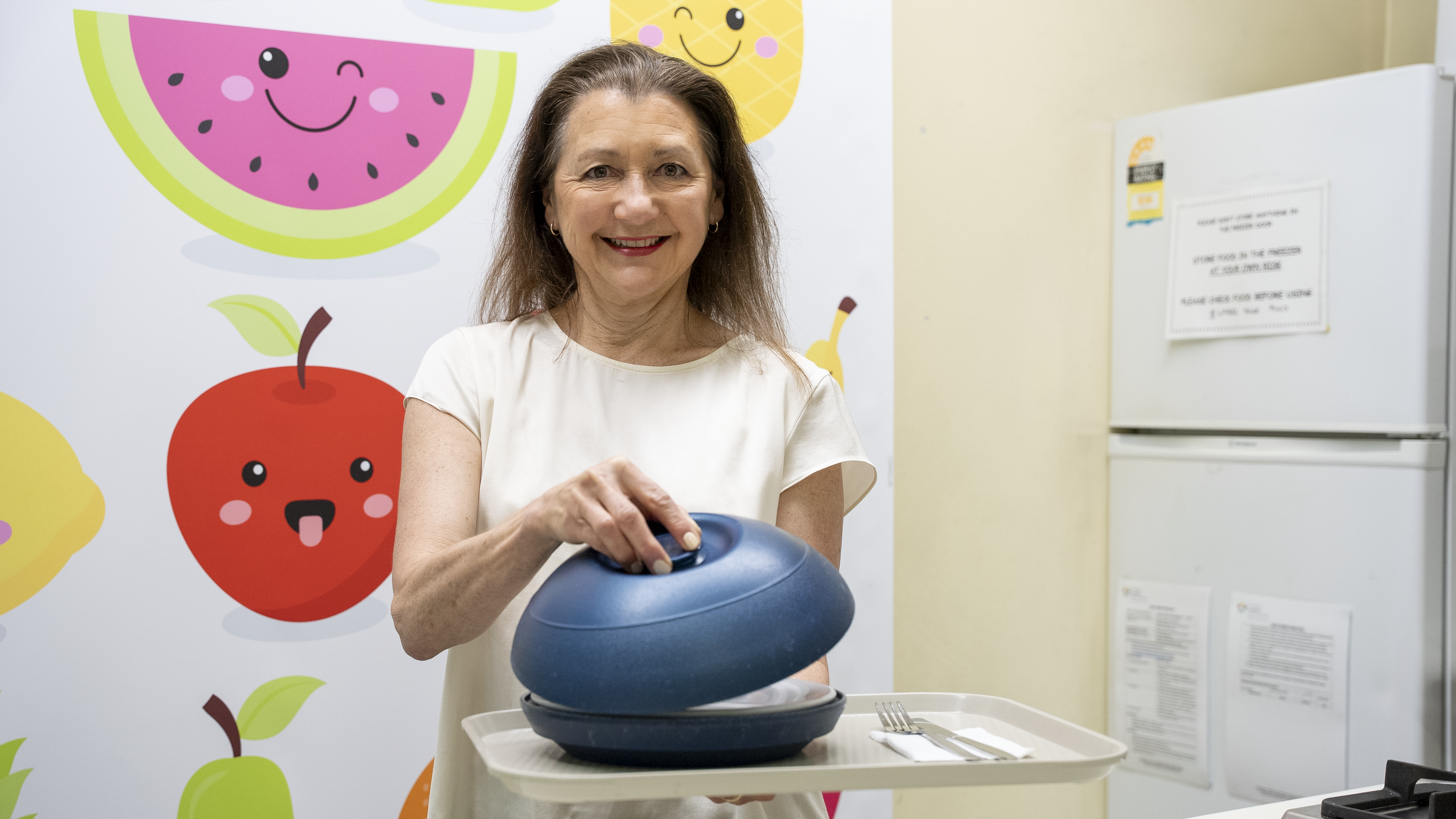 Person holding a food tray with a blue dish cover in a room with colorful fruit illustrations on the wall and a refrigerator in the background.