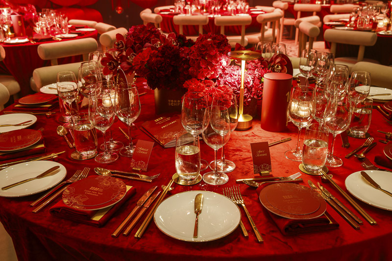 A circular red dining table with shiny gold utencils and bright red flowers and decorations adorned in the centre