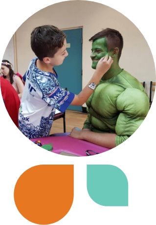 A boy painting a man dressed as the hulk's face with green paint