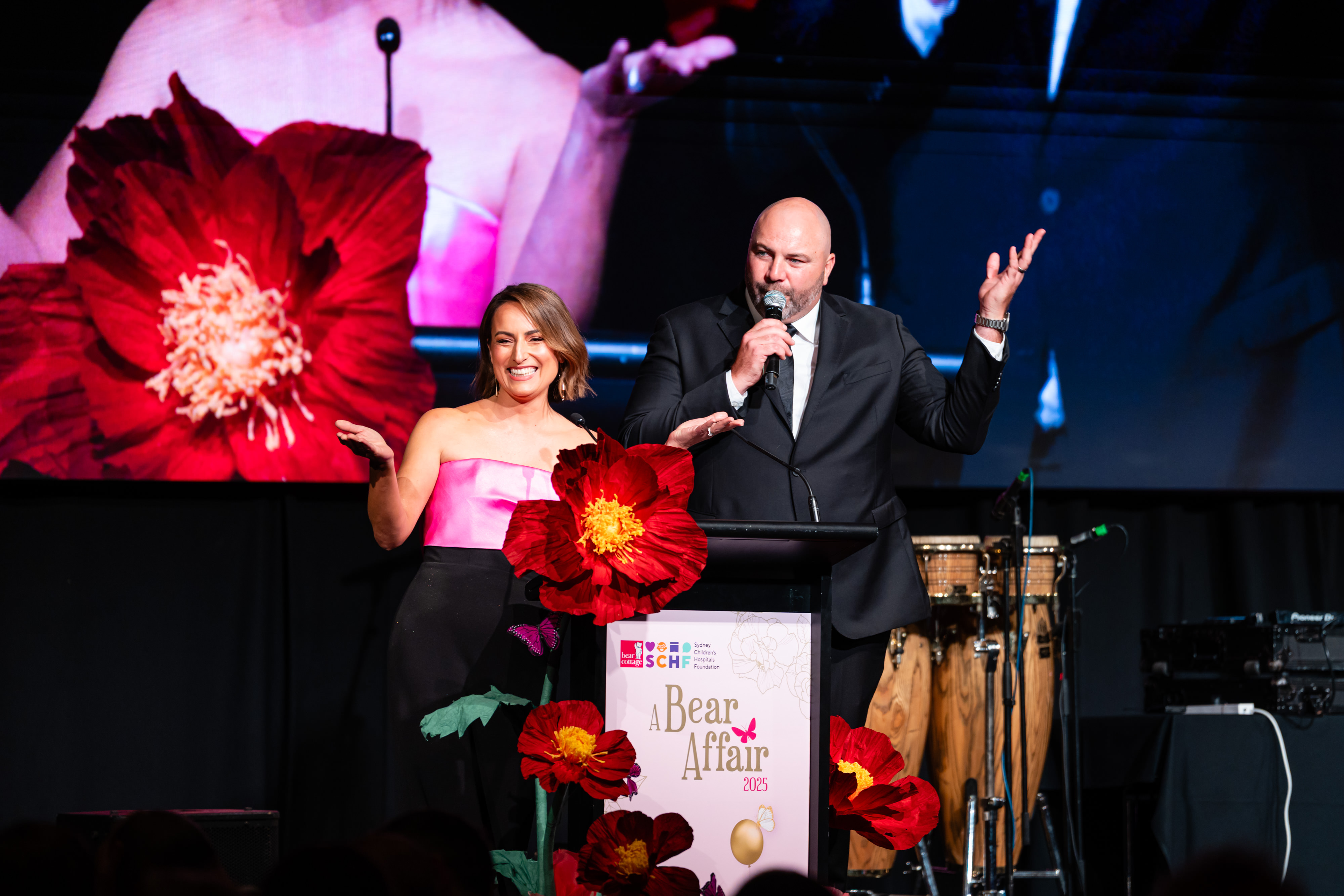 Two presenters speaking at a lectern decorated with oversized red flowers.