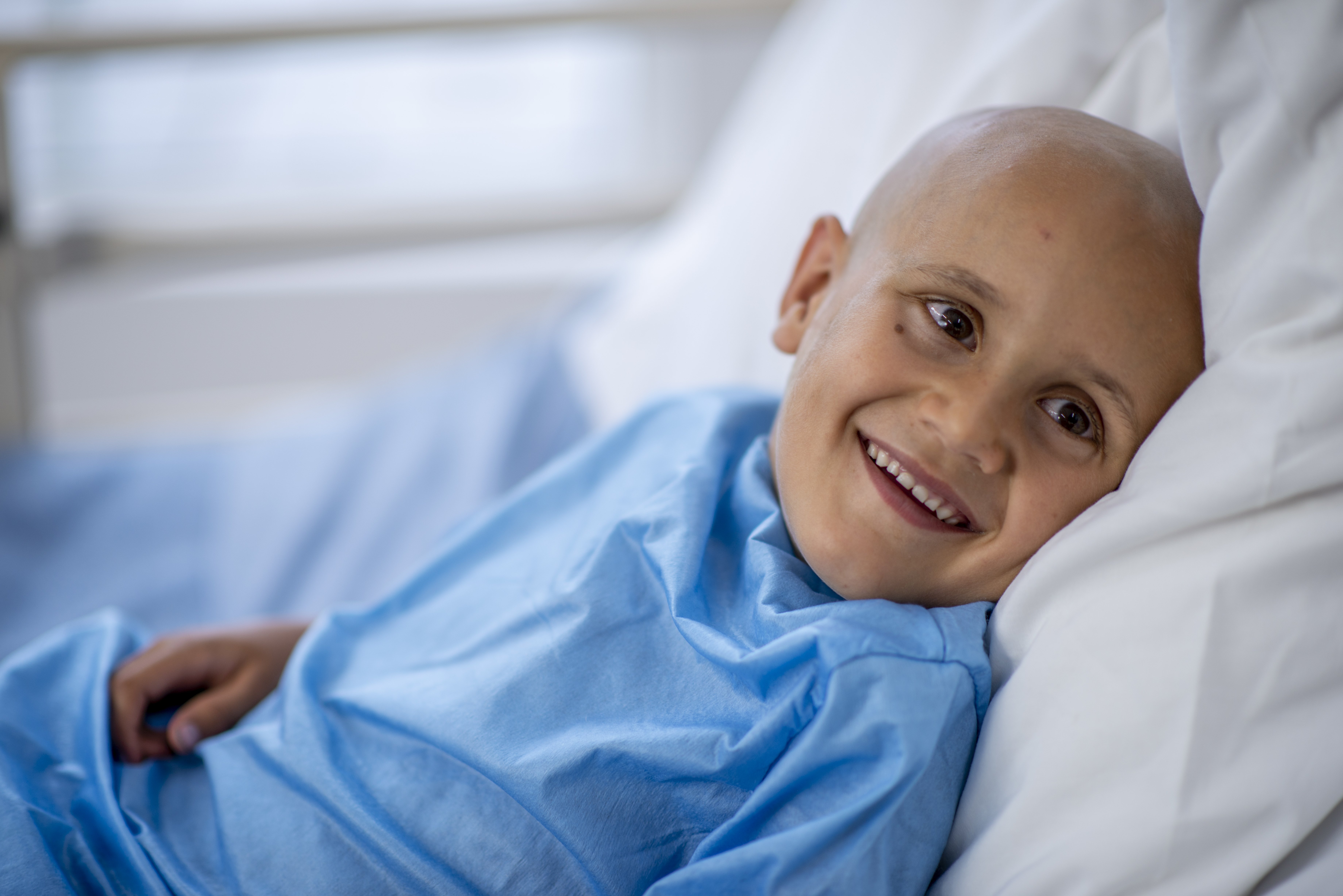 Smiling young patient with a shaved head, wearing a blue hospital gown and resting against white pillows in a bright hospital room