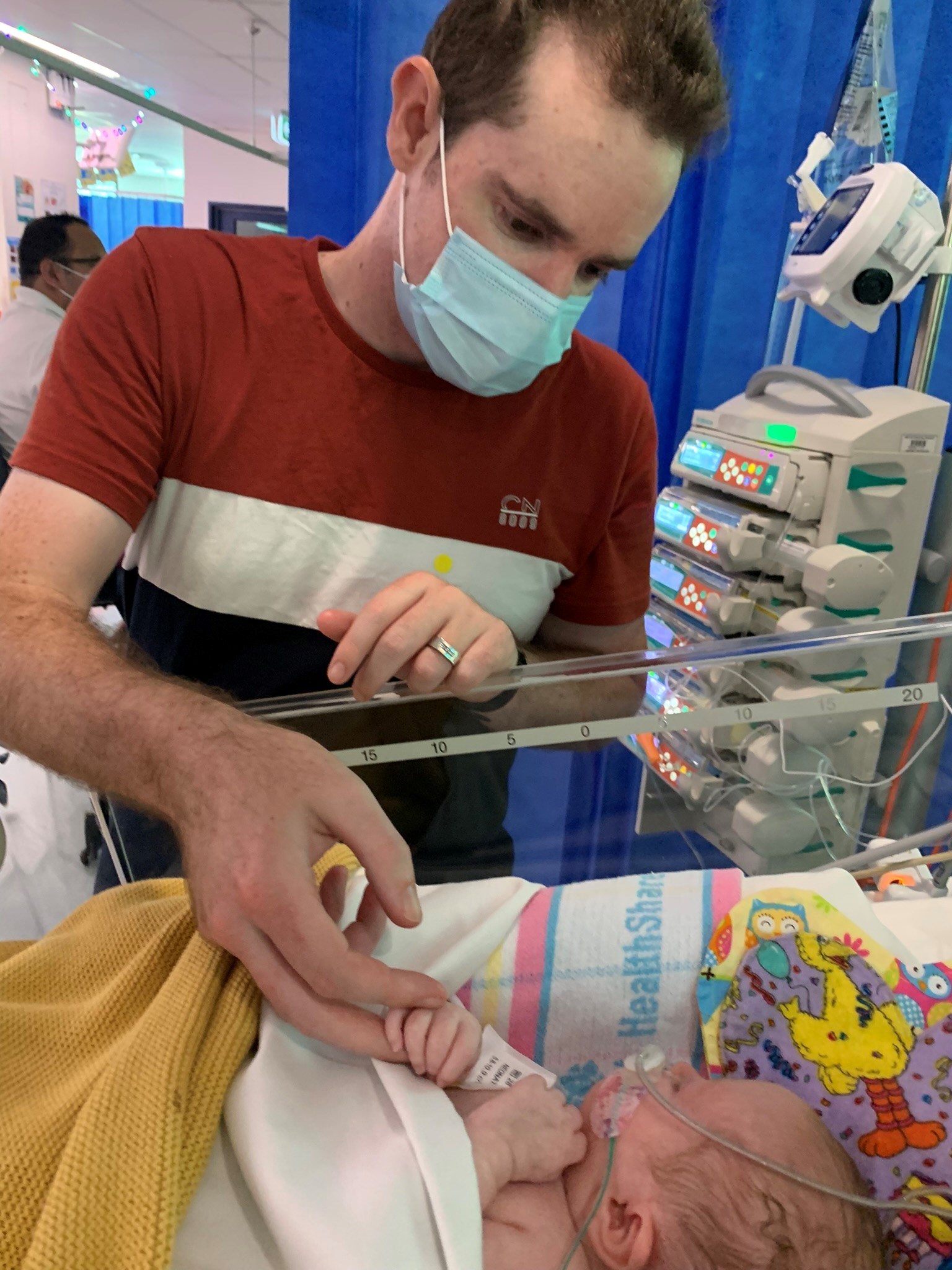 Image of father wearing face mask while holding small baby's hand in crib in hospital.