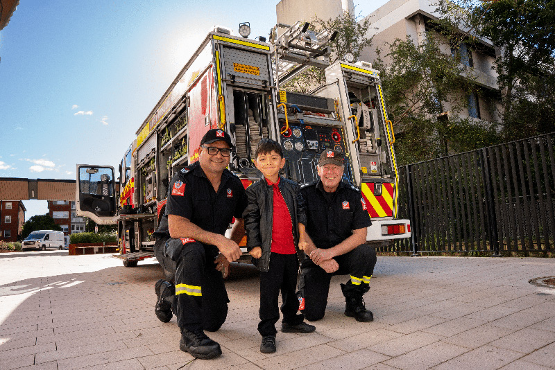 A young boy standing between two firefighters. Behind them is a bright red fire engine.