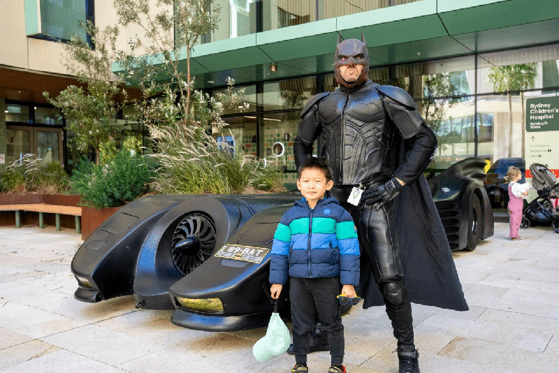 A young boy standing next to a man dressed as Batman outside one of the children's hospitals on Sydney Sick Kids Day. Behind them is a sleek black car that looks like the Batmobile