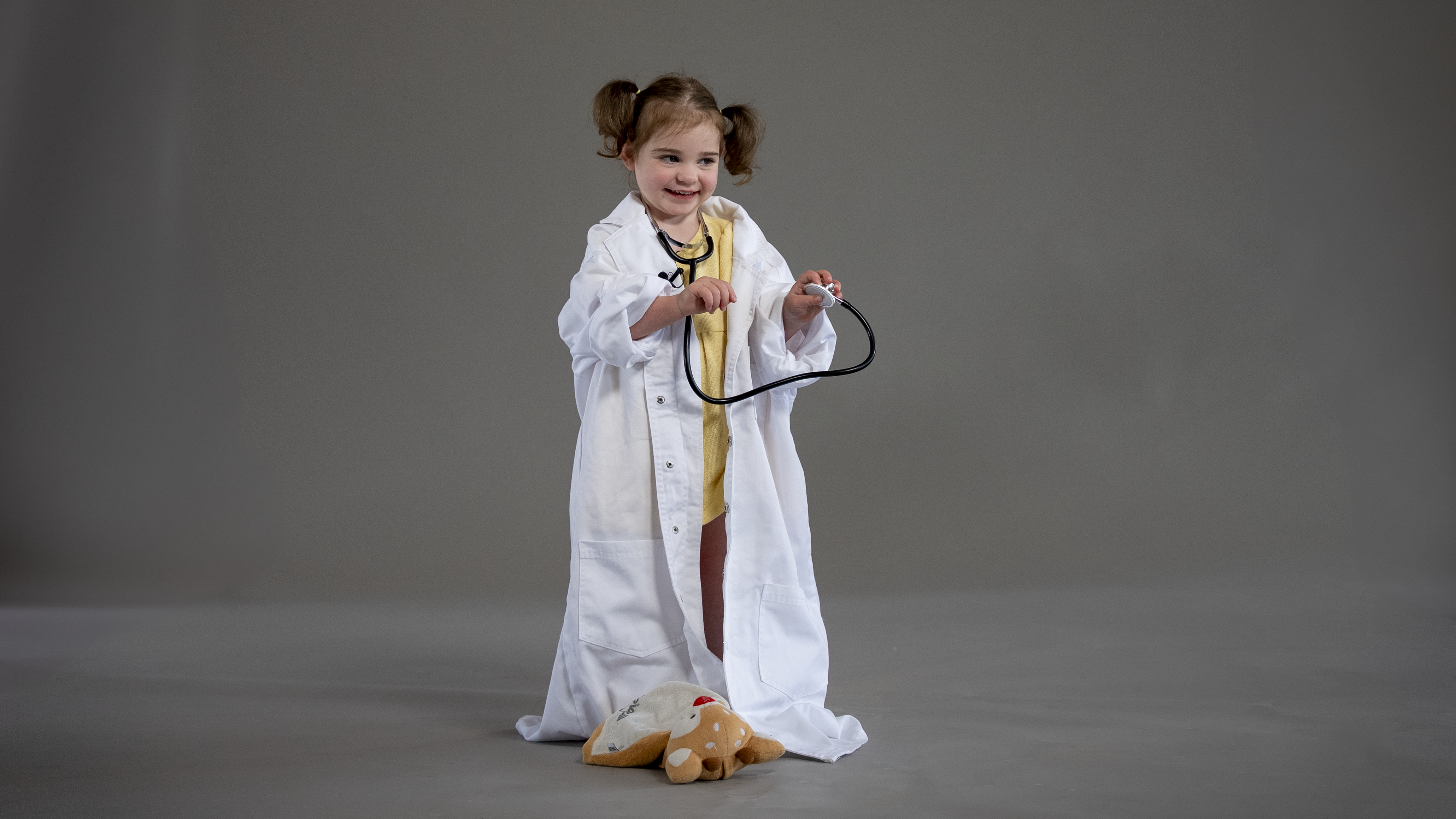 Child wearing an oversized white lab coat and holding a stethoscope, standing on a plain grey background with a plush toy on the floor.