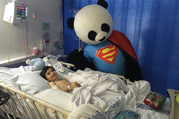 Monty laying in a hospital bed. A person in a panda superhero costume is waving next to him